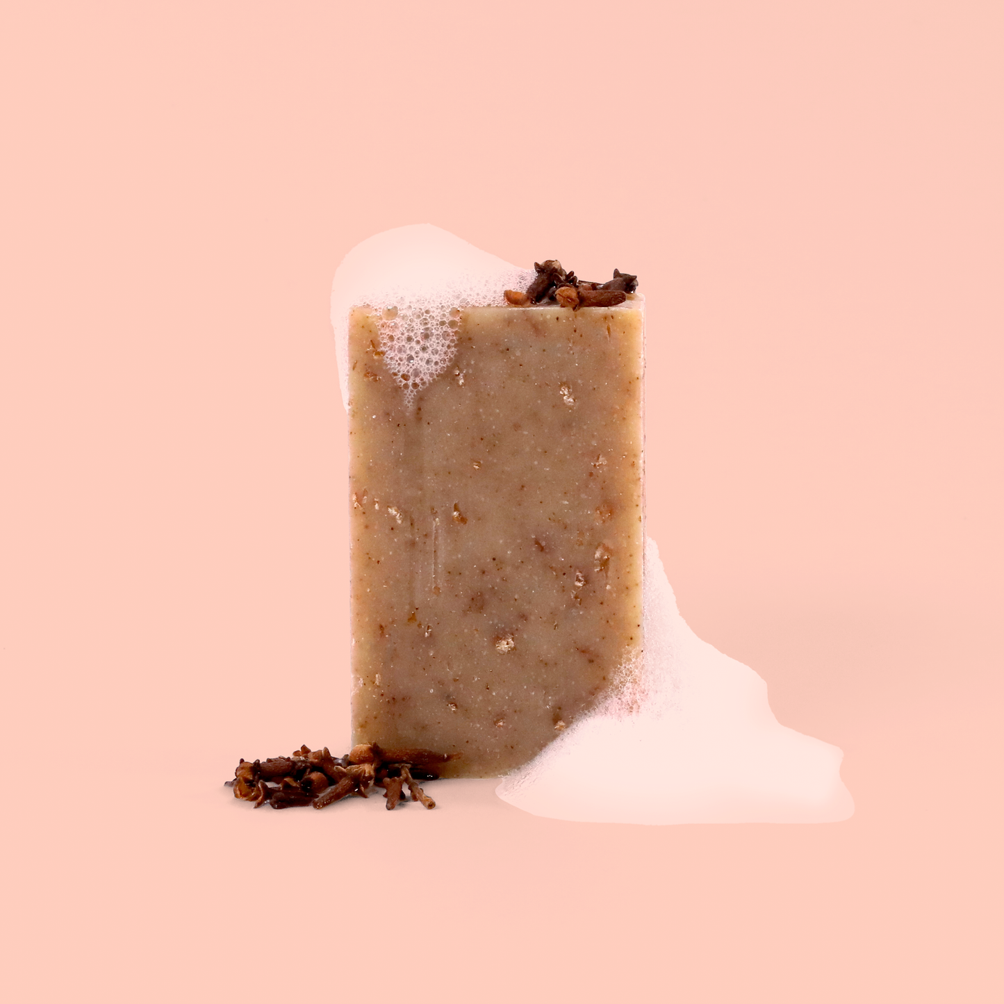 Close-up of a beige speckled soap bar with scattered cloves or spices on top, shown against a pink background with soap bubbles or foam details. The soap appears to have a natural, handmade texture.