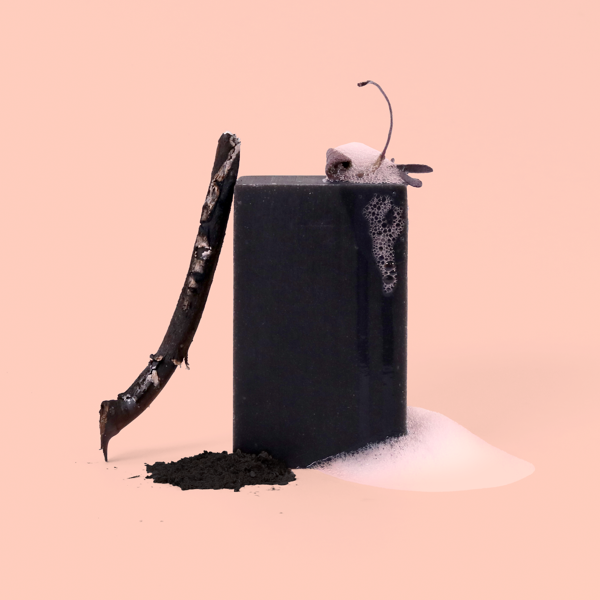 A black bar of soap styled with charcoal dust or powder, a burnt wooden stick, and white foam elements against a pink background. The composition creates a dramatic contrast between the dark soap and light background.