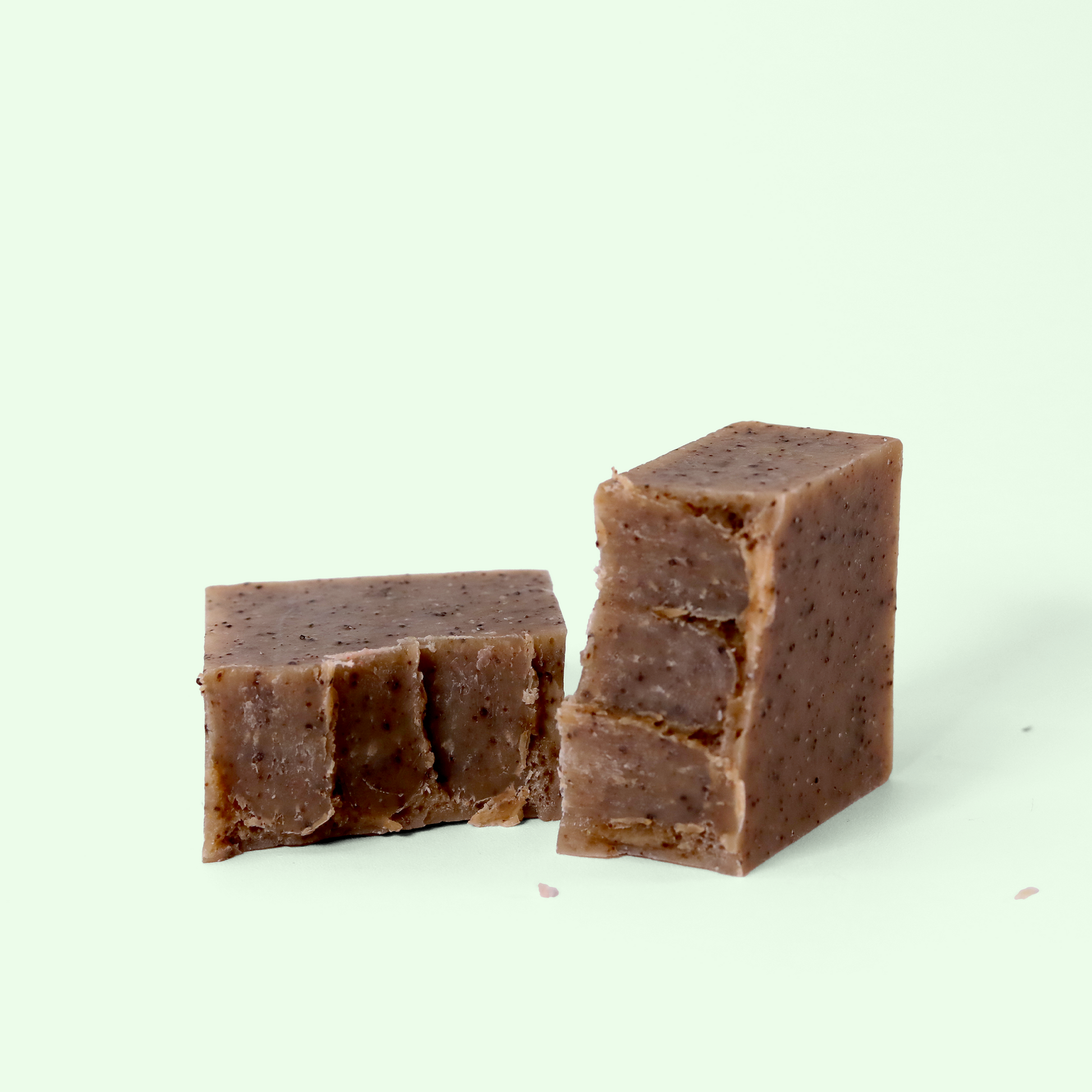 A rectangular block of brown soap with visible specks on a light background, broken in half. Both blocks have a rough, textured surface with small cracks and imperfections visible. Sun Basin Soap - Good Store.