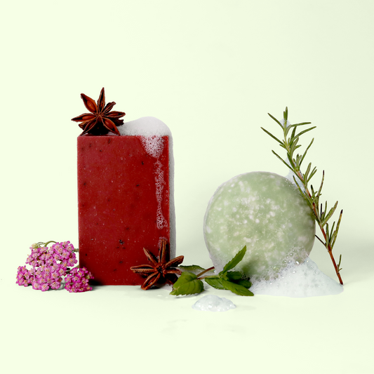 Artisanal soap arrangement featuring a red rectangular soap bar with star anise, and a round pale green shampoo bar with rosemary. Pink yarrow flowers, foam, and herbs decorate the composition, set against a light background. Sun Basin Soap - Good Store. 