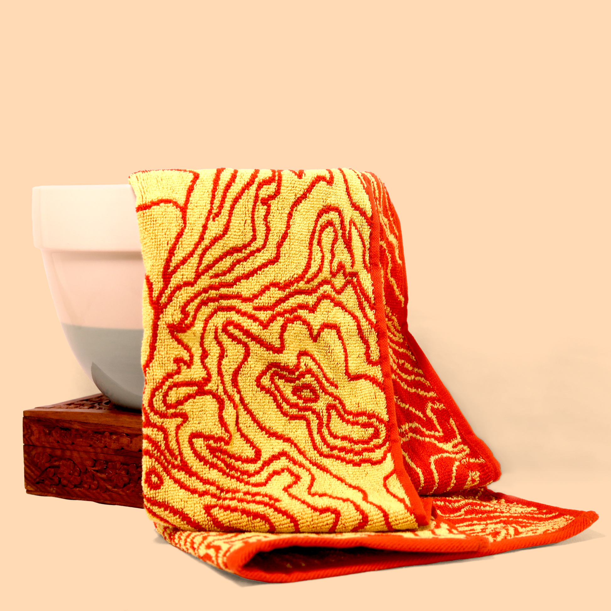 A yellow and red topographic patterned towel draped over a white ceramic bowl and a wooden box. The towel has a bold, abstract design with red lines on a yellow background. The composition is set against a peach-colored background. Sun Basin Soap - Good Store. 