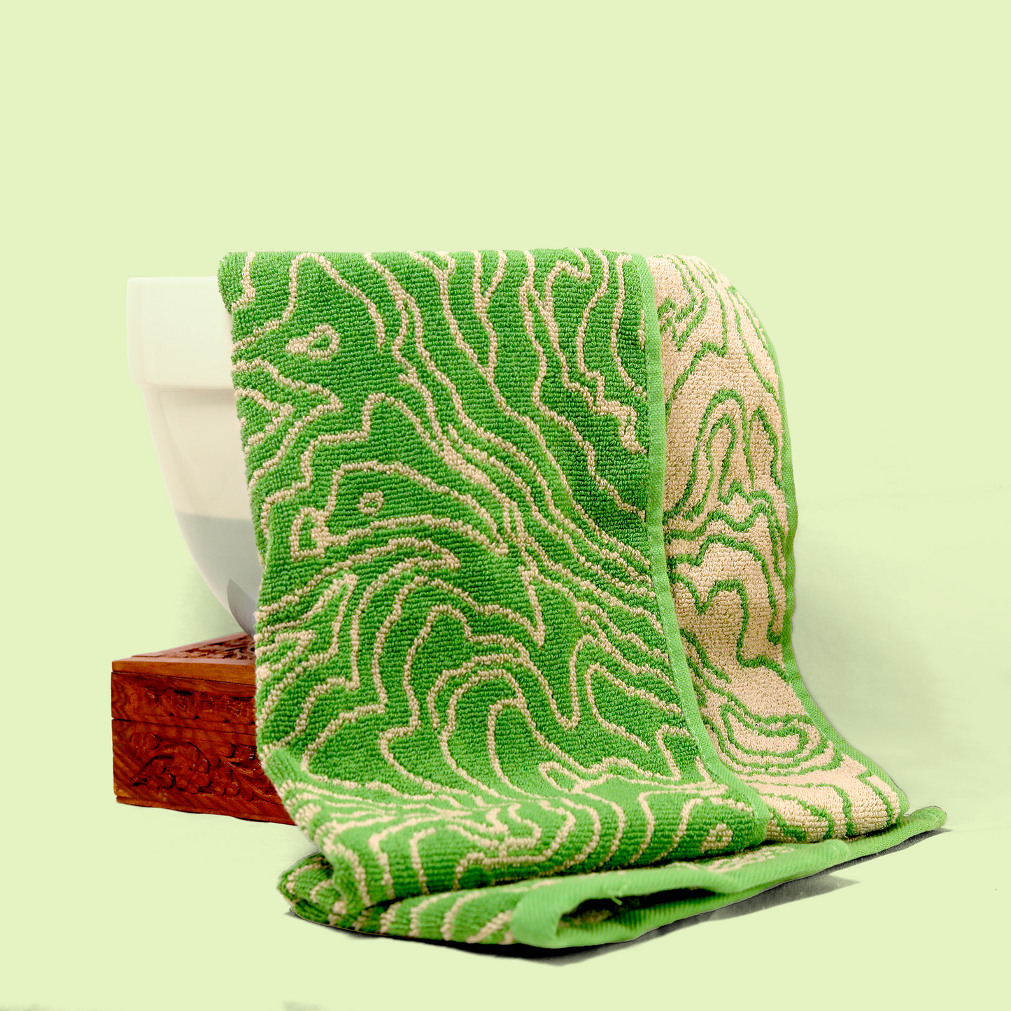 A green and beige topographic patterned towel draped over a white ceramic container and a wooden box. The towel features an intricate swirling design, with beige lines on a green background. Sun Basin Soap - Good Store.