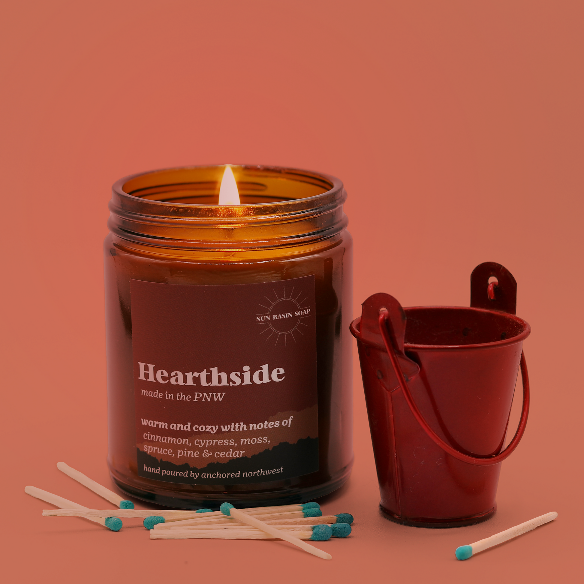 A candle in an amber jar with the text "Hearthside" and the Sun Basin Soap logo sits next to a little red tin, surrounded by matches. A Good Store product. 