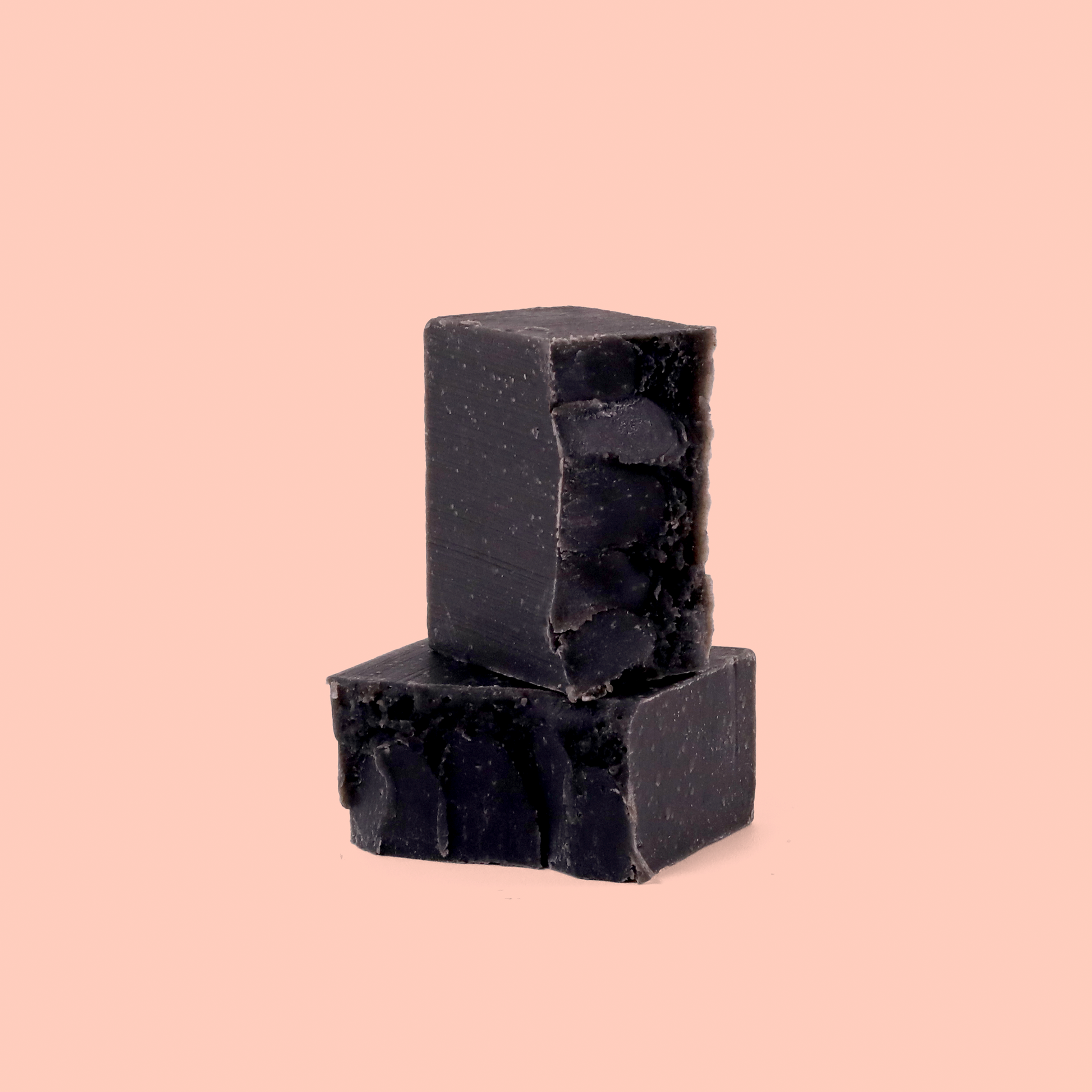 Dark charcoal-colored soap bars stacked on a pale pink background. The soap bars appear to be natural or handmade with a rough, rustic texture.