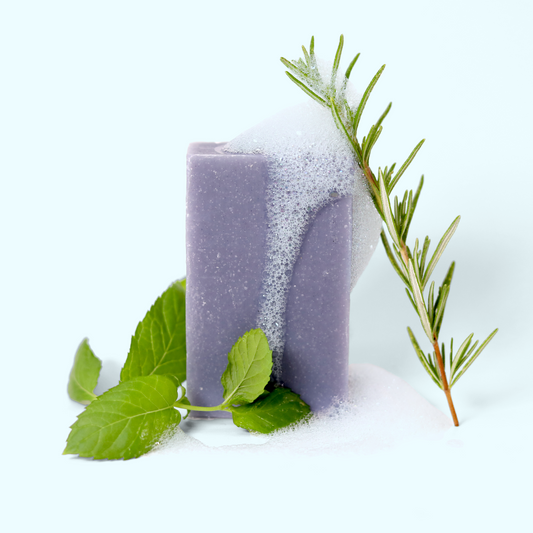 A rectangular block of purple-gray Navigator soap partially covered in white foam or lather. The soap is surrounded by fresh herbs: a sprig of rosemary stands tall to the right, and bright green mint leaves are placed at the base of the soap. The foam appears to be cascading down the side of the soap block. The soap is set against a light background. Sun Basin Soap - Good Store.