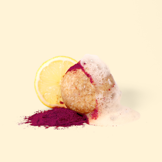 A round, beige shampoo bar partially covered in foam and dusted with bright pink powder. A slice of lemon and a pile of pink powder are placed next to the soap. The composition is set against a cream-colored background. Sun Basin Soap - Good Store.