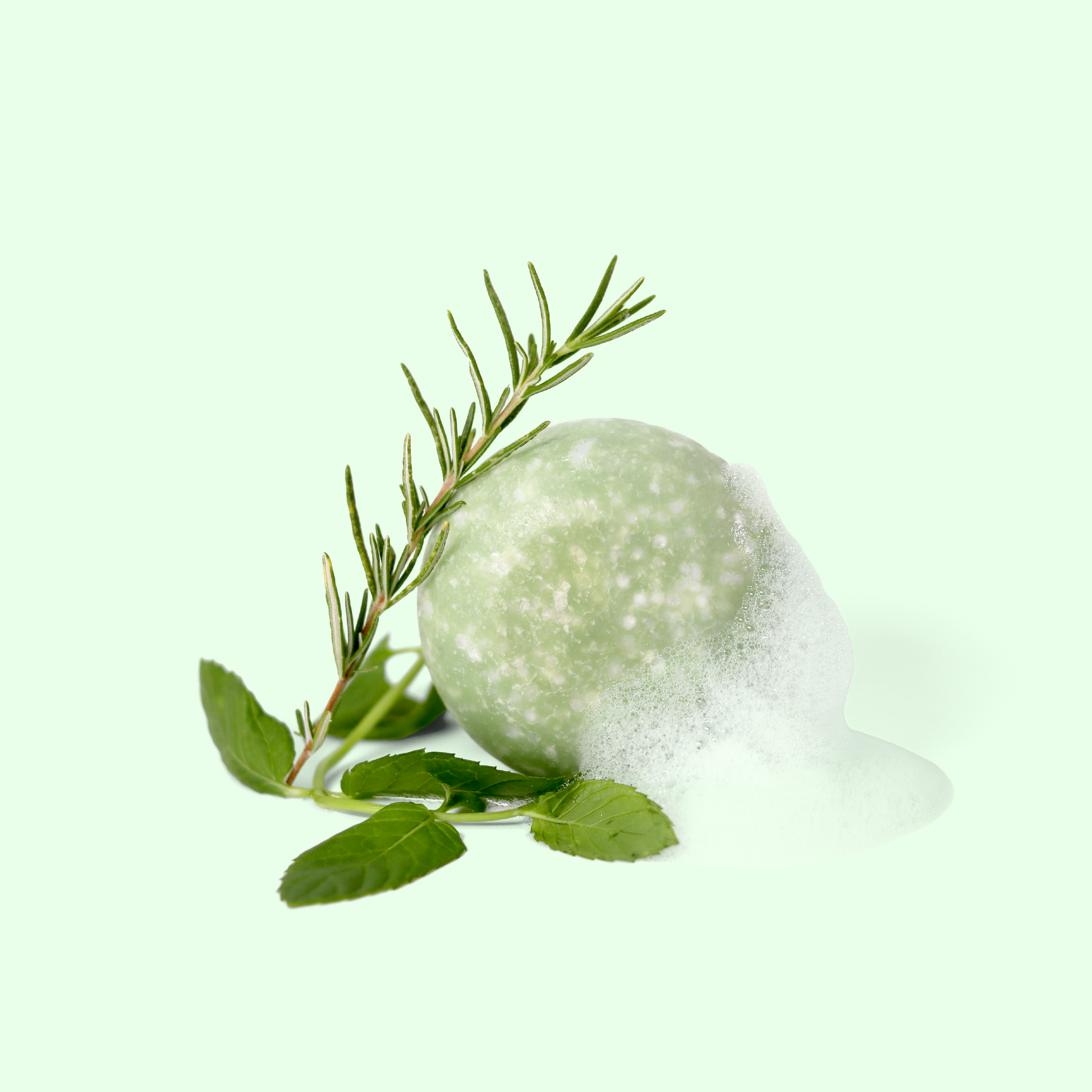 A round, pale green shampoo bar is shown partially covered in foam. A sprig of rosemary is resting on top of the soap, and there are fresh mint leaves placed near the base. Sun Basin Soap - Good Store. 