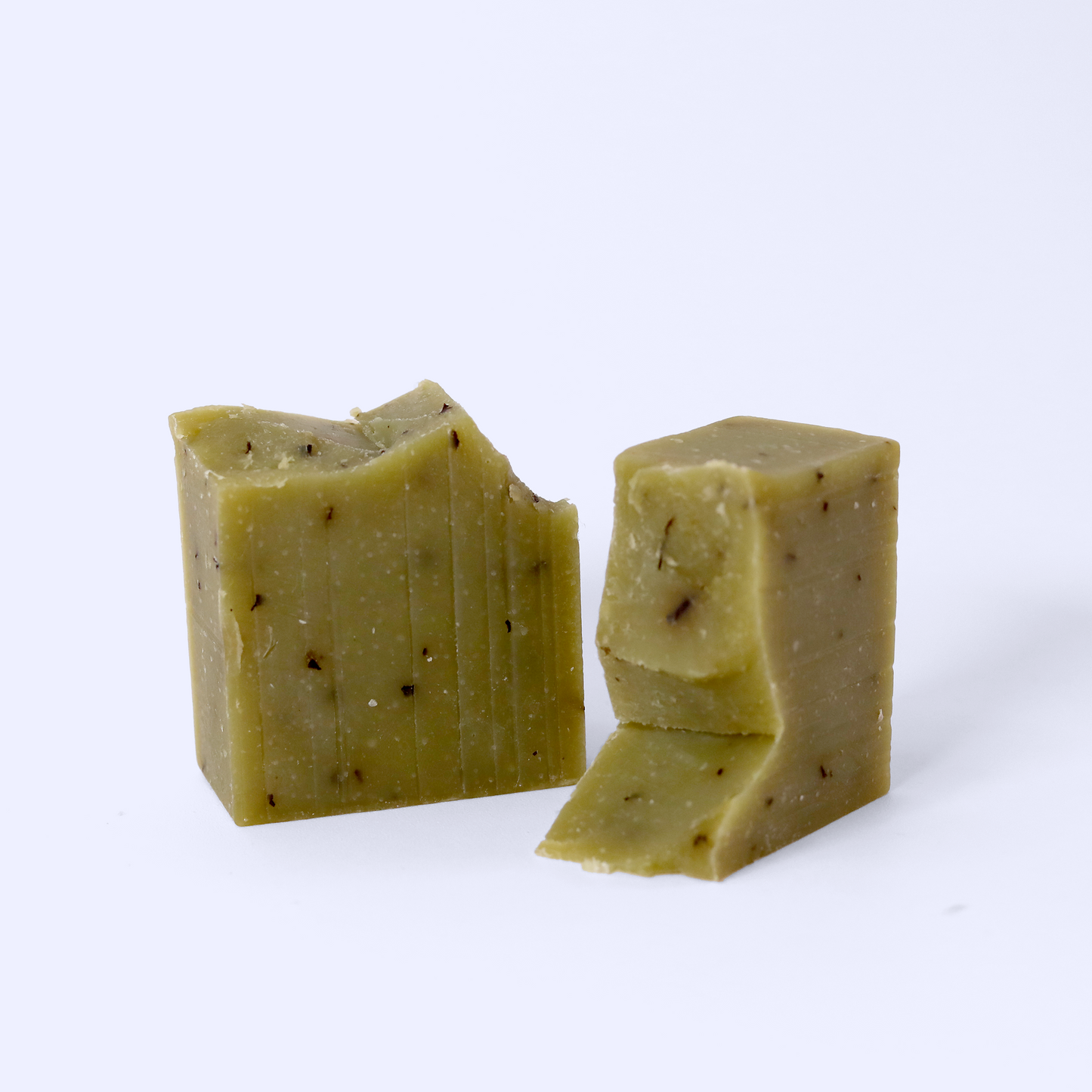 Two pieces of olive-green soap with visible specks. The soaps are photographed on a light background, emphasizing their natural, handmade appearance. Sun Basin Soap - Good Store.