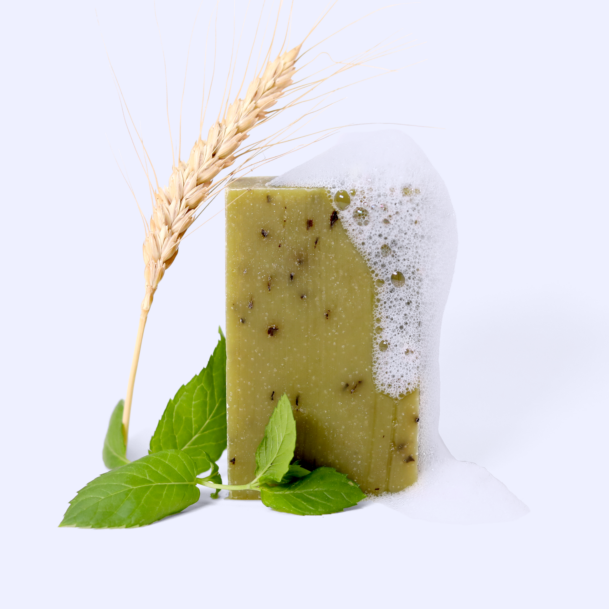 A rectangular bar of green Snowmelt soap with visible natural specks, partially covered in foam. A wheat stalk leans against it, and fresh mint leaves are placed at its base. Sun Basin Soap - Good Store.