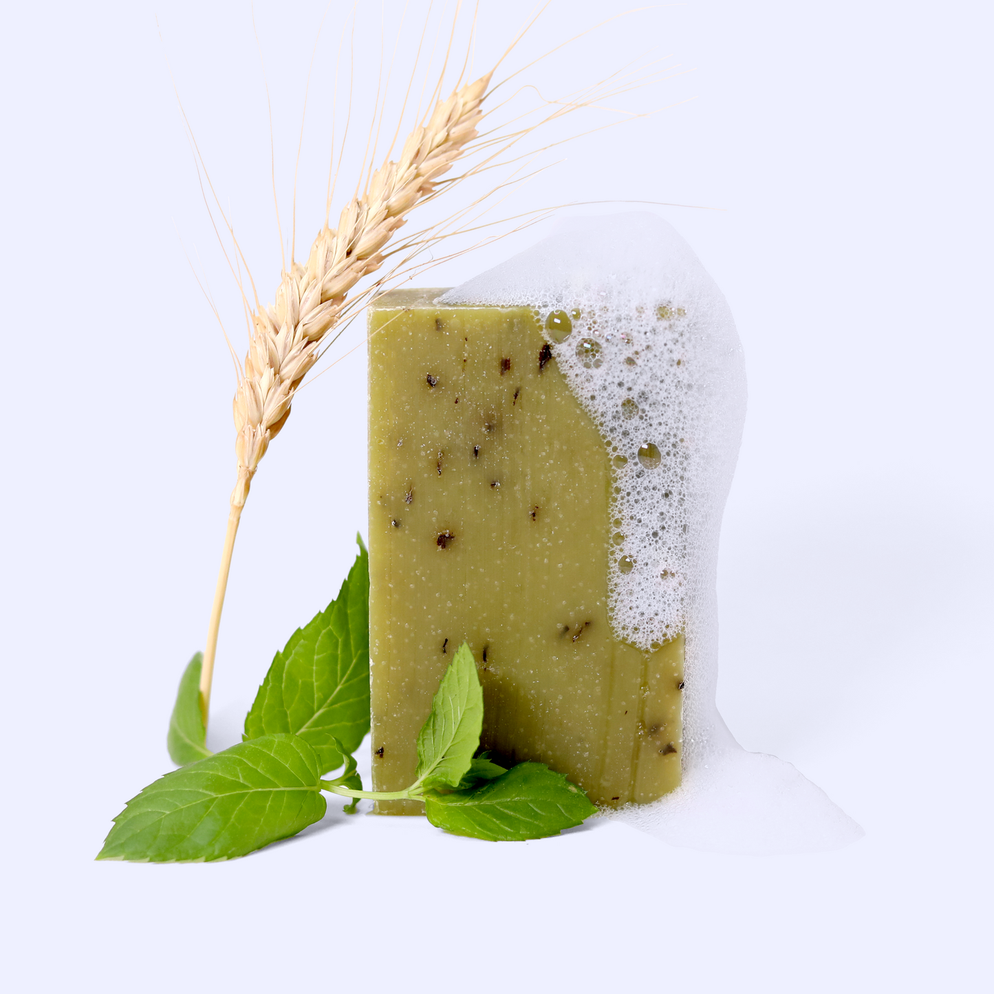 A green bar of soap with visible botanical specks embedded in it, partially covered with white foam or lather. Fresh mint leaves are positioned at the base of the soap, and a stalk of wheat or grain is placed alongside it. The soap has a natural, herbaceous appearance and is photographed against a light blue background.