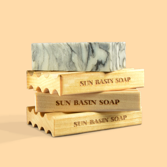  A stack of soap savers from Sun Basin Soap. On top is a marbled gray bar of soap, sitting on three wooden soap dishes. The wooden dishes have wavy edges and are engraved with "SUN BASIN SOAP". The dishes are in varying shades of light wood. The arrangement is set against a pale peach background. 