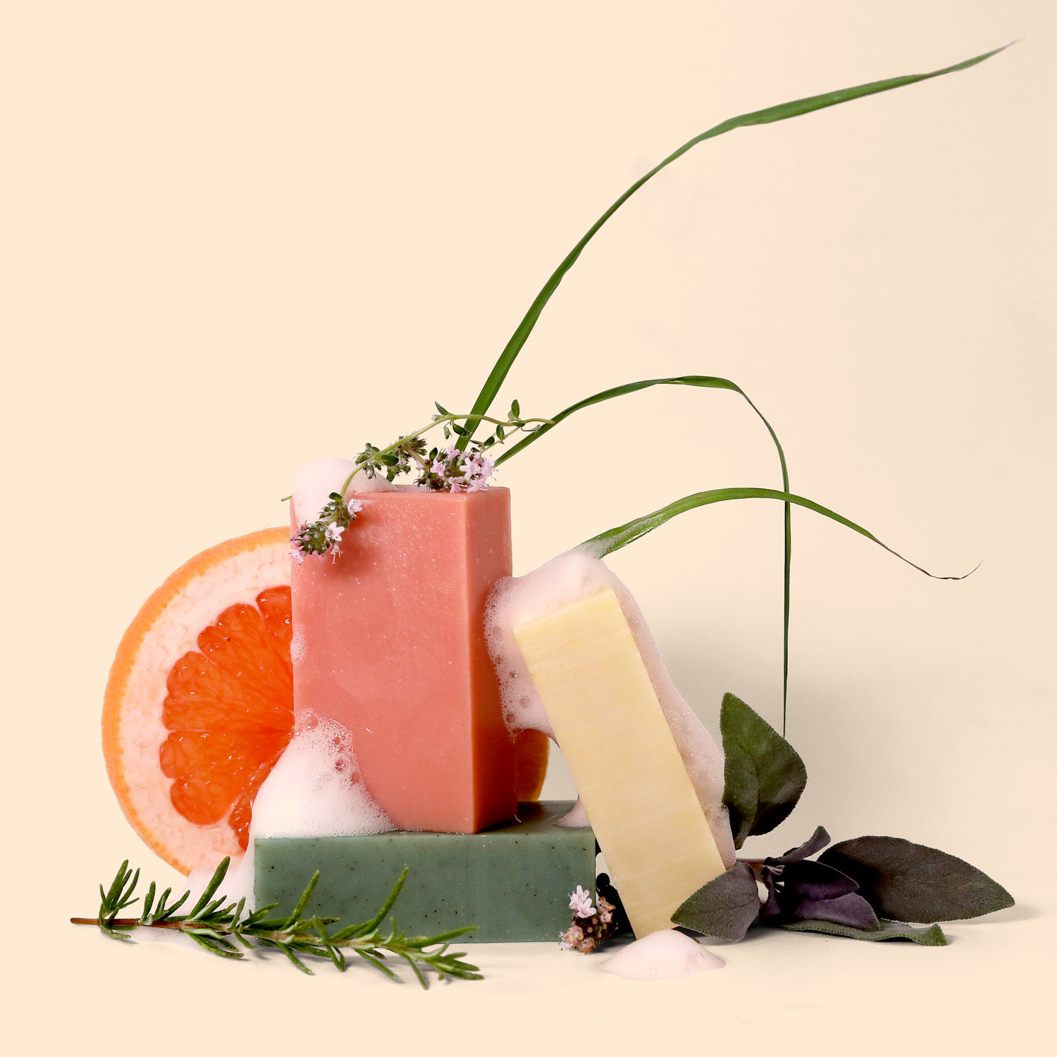 Arrangement of handmade soap bars in pink, pale yellow, and green, with a slice of grapefruit, rosemary sprig, grass-like leaves, and purple sage leaves. White foam partially covers the soaps. Set against a light beige background. Sun Basin Soap - Good Store.