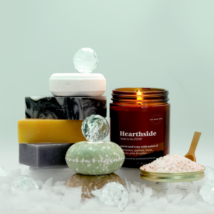 The Viridian Sun Bath Box from Sun Basin Soap contains a stack of three soap bars with a bath bomb resting atop. Two shampoo bars sit in front of a candle and bath salts. A Good Store product. 