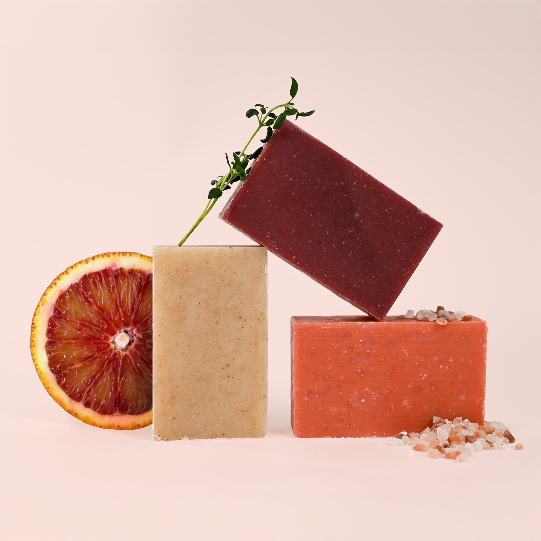 Three handmade soap bars artfully arranged against a pink background - a speckled beige bar, a burgundy bar balanced on top with a plant sprig, and a coral-orange bar with scattered salt crystals. A halved blood orange slice completes the natural skincare product composition.