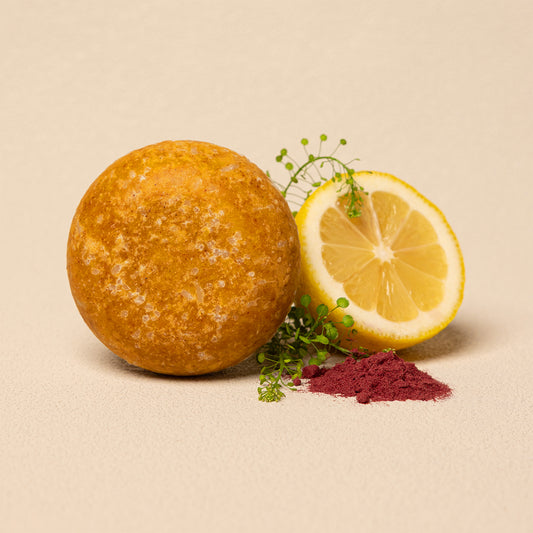 A round, textured golden-brown shampoo bar sits next to a halved lemon and a small pile of deep magenta powder on a warm beige background.