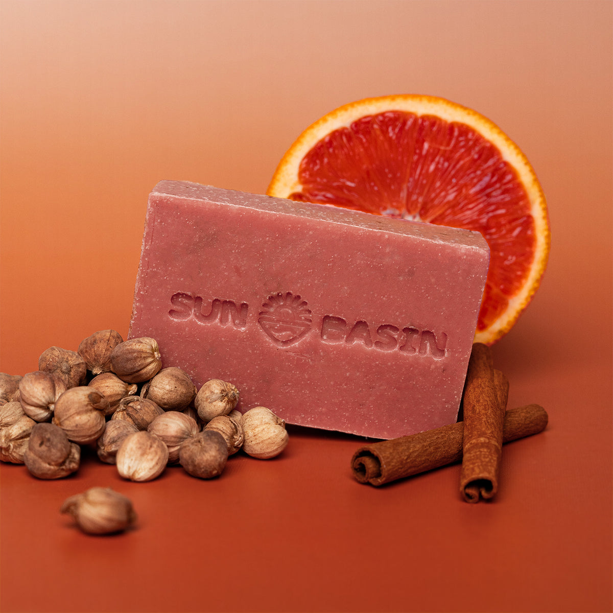 A rectangular bar of Sun Basin soap in a dusty rose color, styled with a sliced blood orange, cinnamon sticks, and whole spices on a warm orange-brown gradient background.
