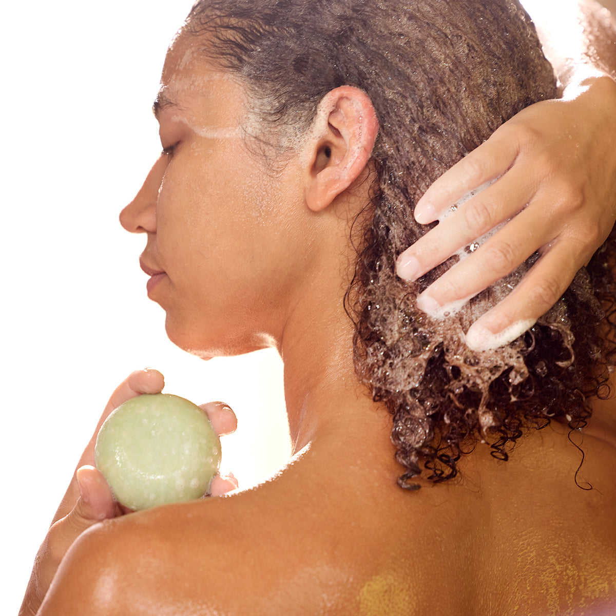 A close-up lifestyle shot of a person with curly dark hair washing their hair, holding a light green shampoo bar in one hand while another hand lathers the hair with white shampoo foam against a bright white background.