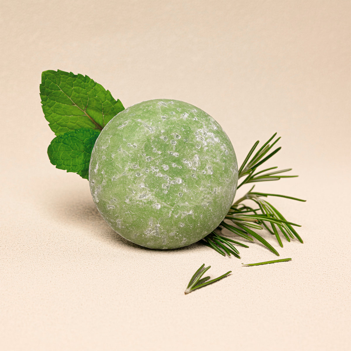 A round green shampoo bar styled with fresh green mint leaves and fresh rosemary on a warm beige background.