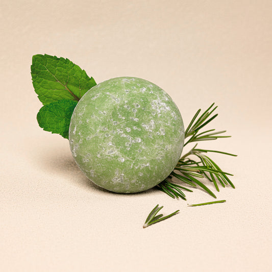 A round green shampoo bar styled with fresh green mint leaves and fresh rosemary on a warm beige background.