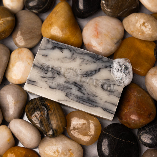 A rectangular handmade soap bar with a white and gray marbled pattern and a small white bead accent, styled on a bed of smooth, polished river stones.