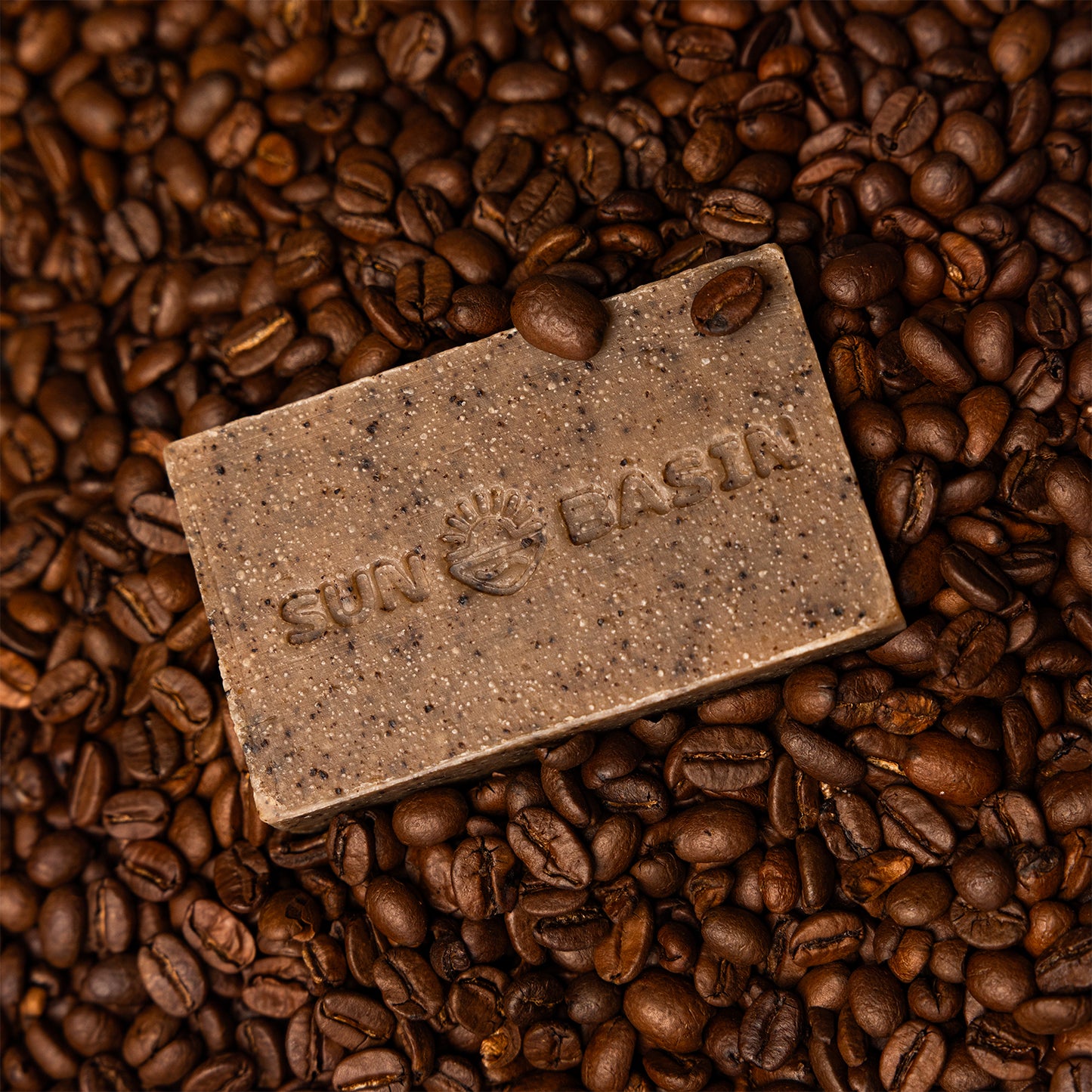 A rectangular bar of soap in a speckled brown color, embossed with the brand name and logo, resting on a bed of roasted coffee beans.
