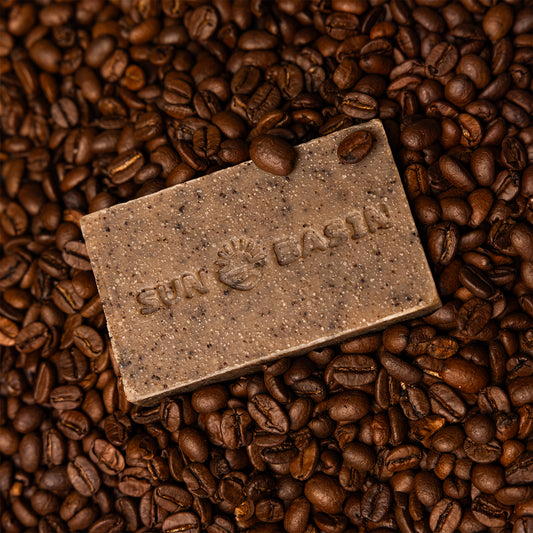 A rectangular bar of soap in a speckled brown color, embossed with the brand name and logo, resting on a bed of roasted coffee beans.