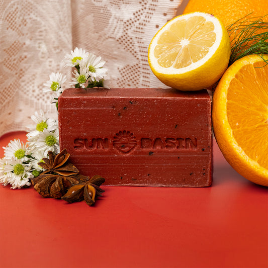 A rectangular bar of Sunbasin soap in a reddish-brown color, embossed with the brand name and logo. The soap is styled on a red surface with fresh lemon and orange slices, white daisies, and star anise, with a lace curtain in the background.