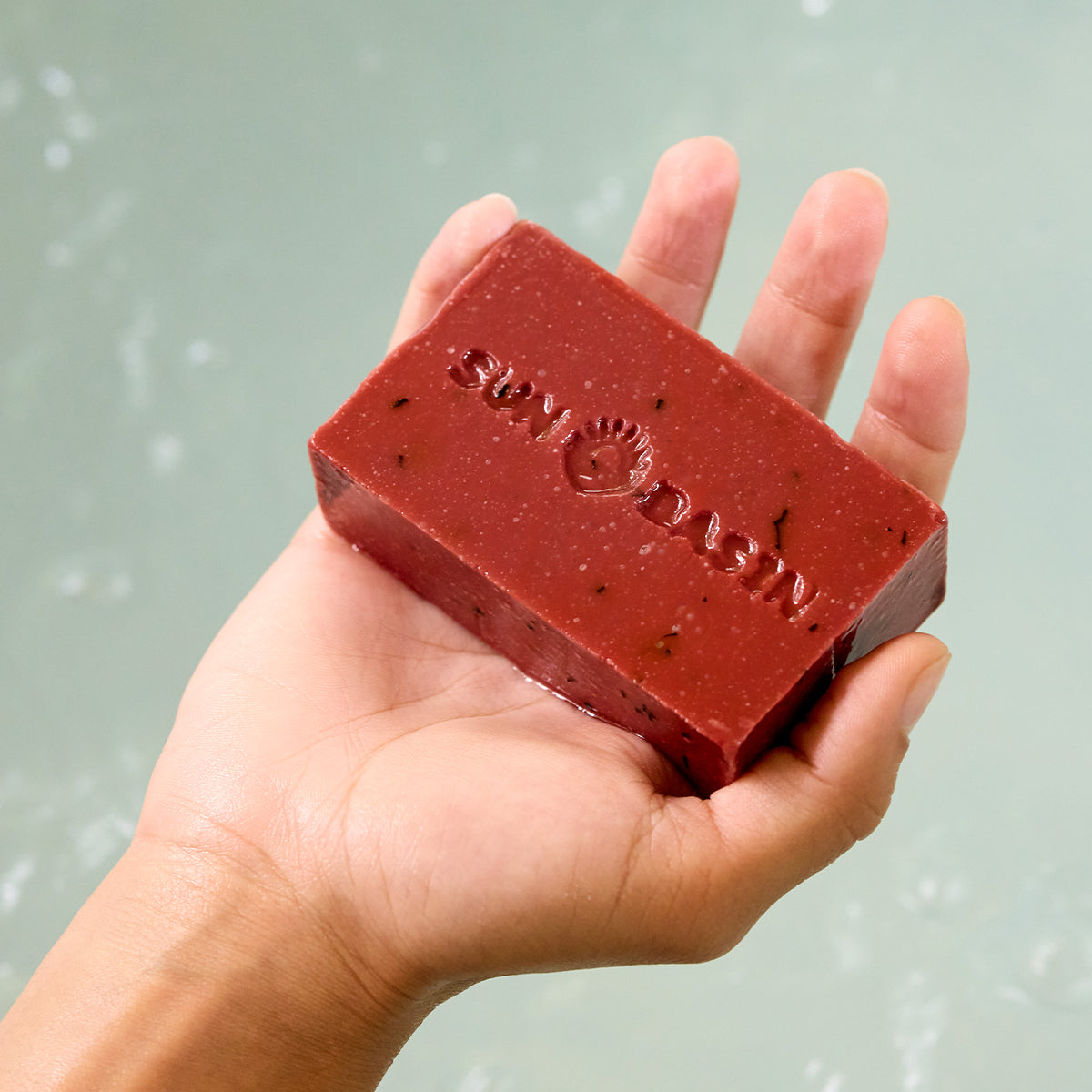 A hand holding a rectangular red soap bar embossed with the brand name 'SEV BASIN' against a soft, pale greenish-blue water background.