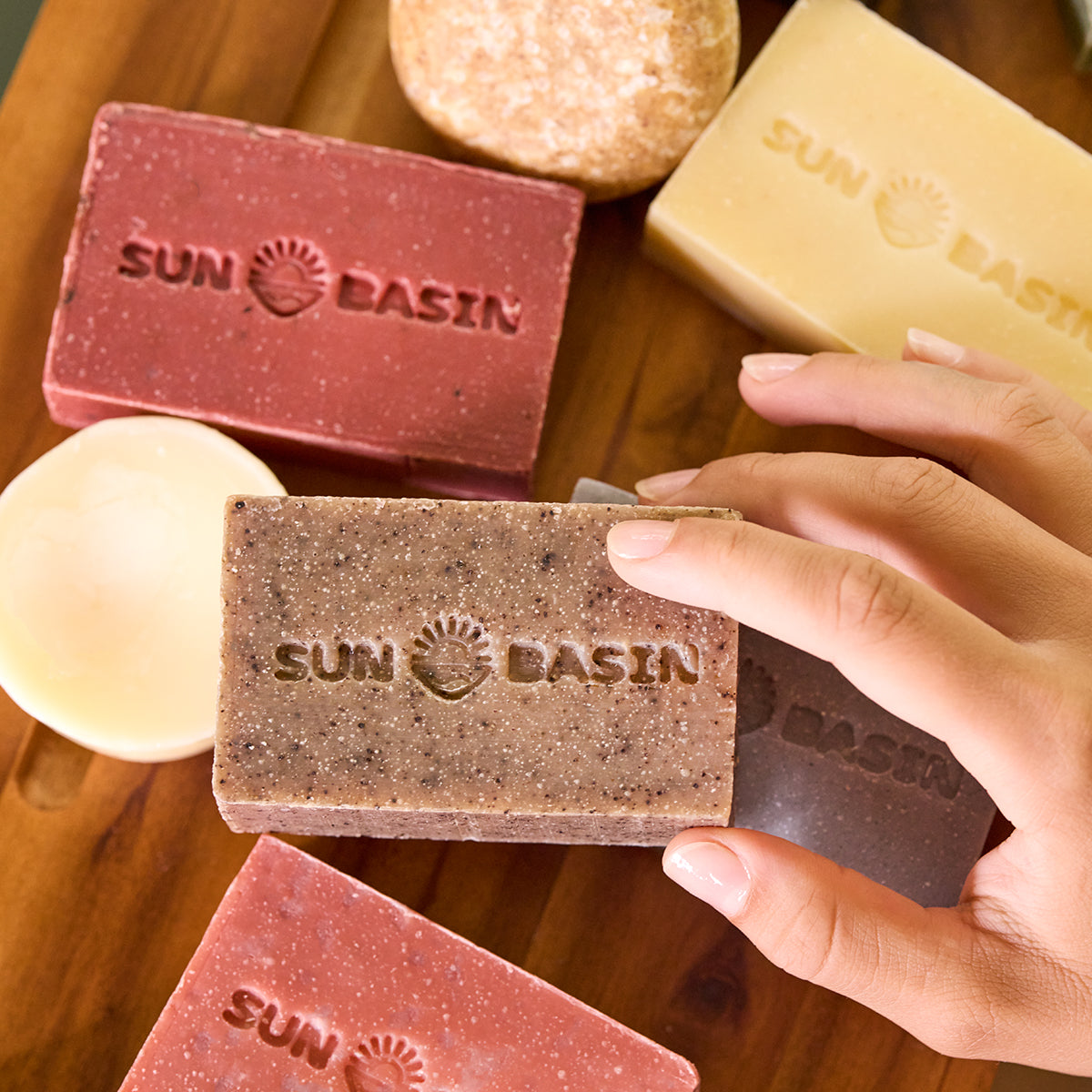 A hand holds a rectangular Sun Basin soap bar in a medium brown shade, surrounded by other colorful soap bars on a wooden surface.