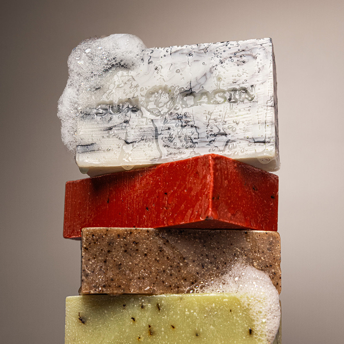 A stacked set of five artisanal handmade soap bars in white, red, brown, and pale yellow, with one bar topped with lathered foam showing the embossed brand name 'Sun basin'.