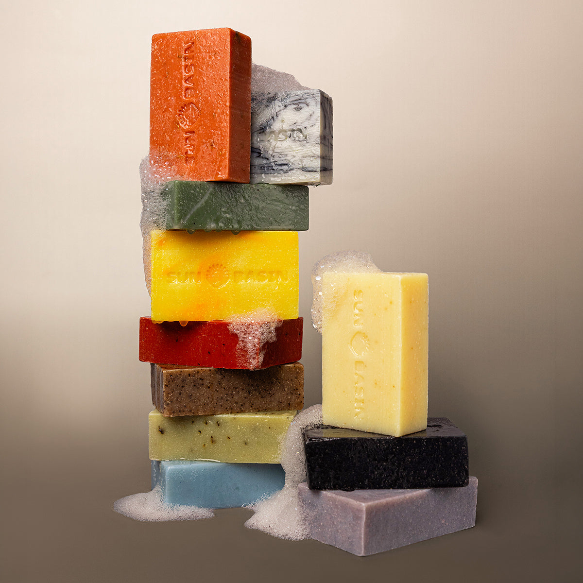 A styled product shot of multiple artisanal soap bars stacked and arranged on a neutral beige background. The soaps come in a variety of colors including orange, green, yellow, red, brown, light blue, black, and lavender, with some showing white lather foam at the top and bottom.