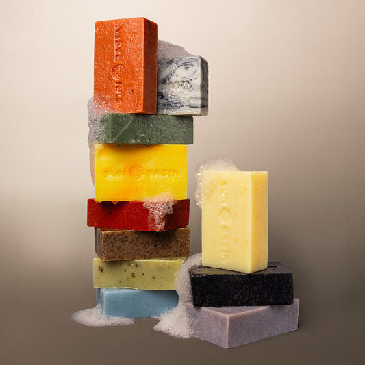 A styled product shot of multiple artisanal soap bars stacked and arranged on a neutral beige background. The soaps come in a variety of colors including orange, green, yellow, red, brown, light blue, black, and lavender, with some showing white lather foam at the top and bottom.