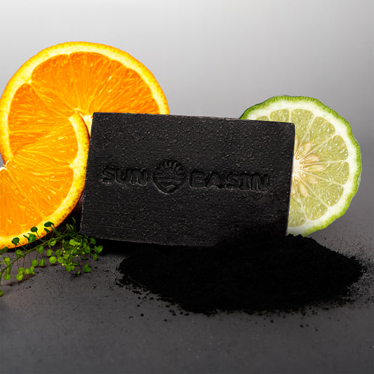 A rectangular black charcoal soap bar with the brand embossed on it, styled with sliced orange, lime, and a small pile of loose black powder on a dark gray surface against a light gray background.