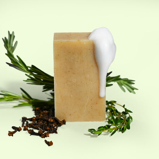 Bar of soap with a white foam, surrounded by green leaves and black cloves on a light background