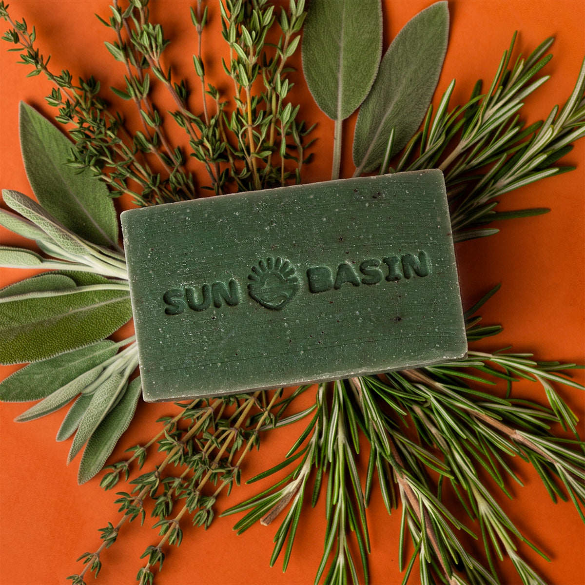 A rectangular green soap bar embossed with 'SUN BASIN' and a sun logo, arranged on an orange background with fresh rosemary, sage, and thyme herbs.