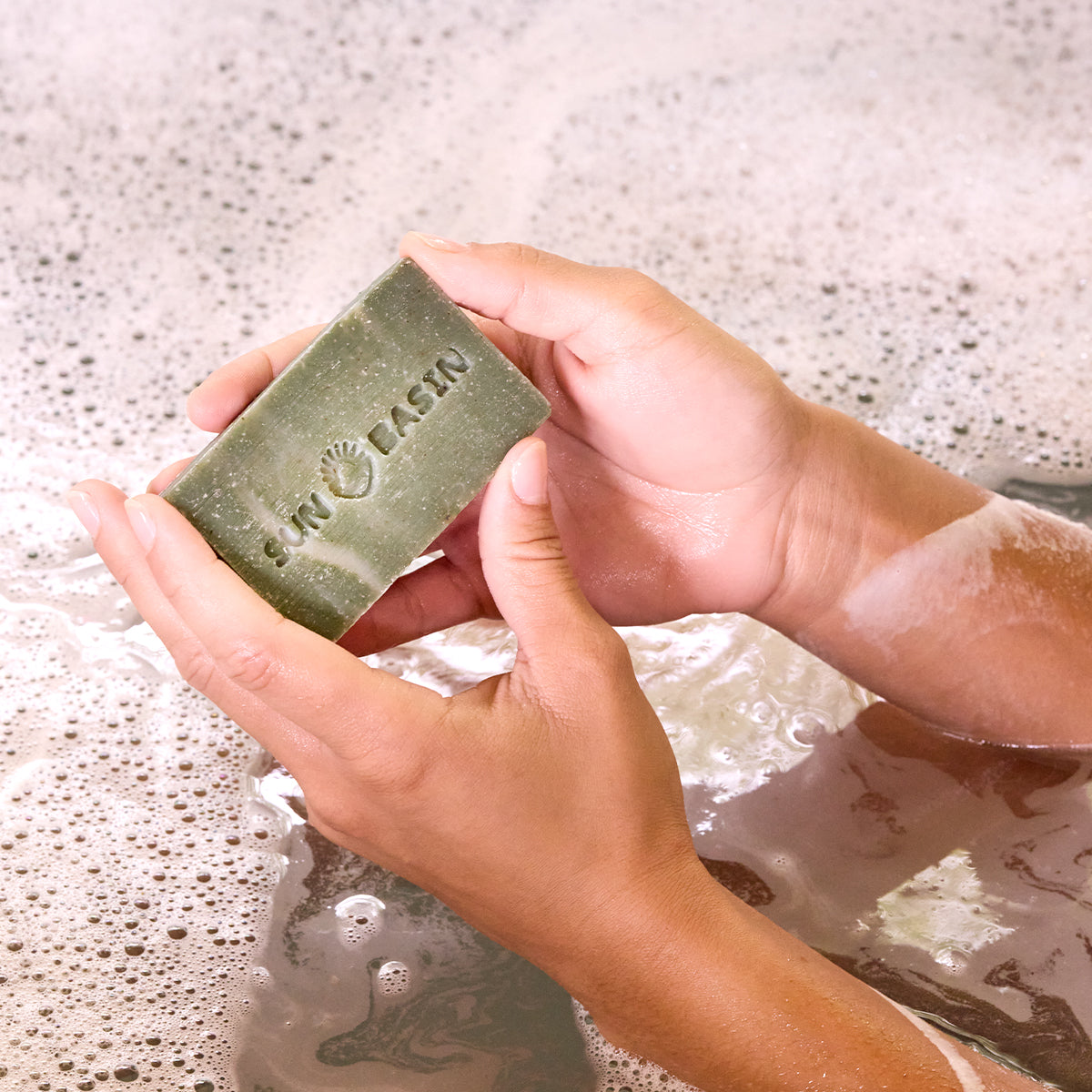 A person holds a rectangular green soap bar labeled 'Sun Basin' in soapy water with foam bubbles around it.