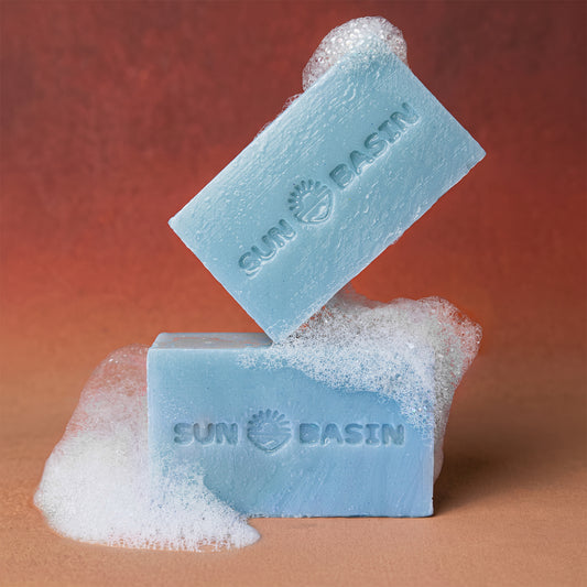 Two light blue Sun Basin soap bars with white foam, one standing upright and the other leaning on top, set against a warm brown gradient background.