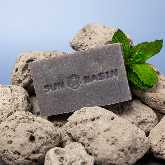 A rectangular bar of gray soap with the brand name 'SUN BASIN' embossed on the front, styled on a bed of light gray volcanic rocks with a green mint leaf on top, against a soft blue gradient background.