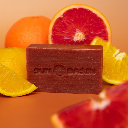 A rectangular bar of Sun Basin soap in a dark reddish-brown color, styled with citrus fruits including oranges, blood oranges, and yellow slices on a warm orange background.
