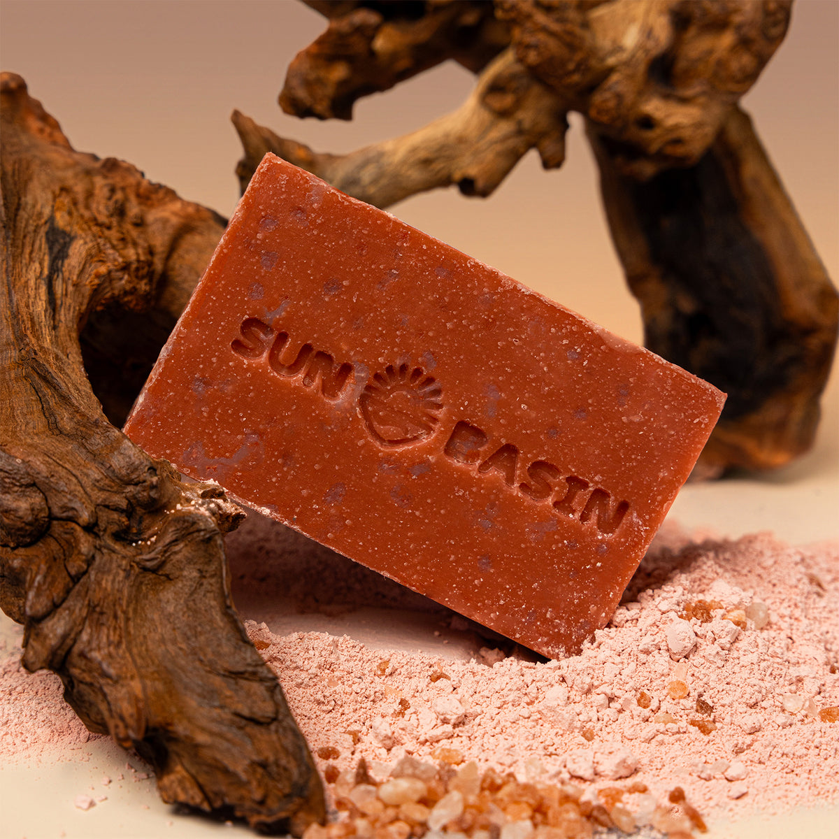 A rectangular bar of terracotta-colored bath salt soap by Sunbath, embossed with the brand name and sun logo, resting on a bed of pink salt crystals next to driftwood.
