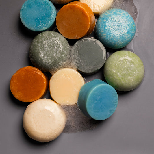 A group of round, disc-shaped shampoo and conditioner bars in assorted colors, including blue, orange, green, cream, and gray, arranged on a dark gray surface with a small amount of water visible between them.
