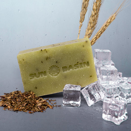 A rectangular bar of Sun Basin soap in a muted yellow-green color, embossed with the brand logo. The soap is styled with clear ice cubes, dried botanicals, and wheat stalks on a gray surface against a light blue-gray background.