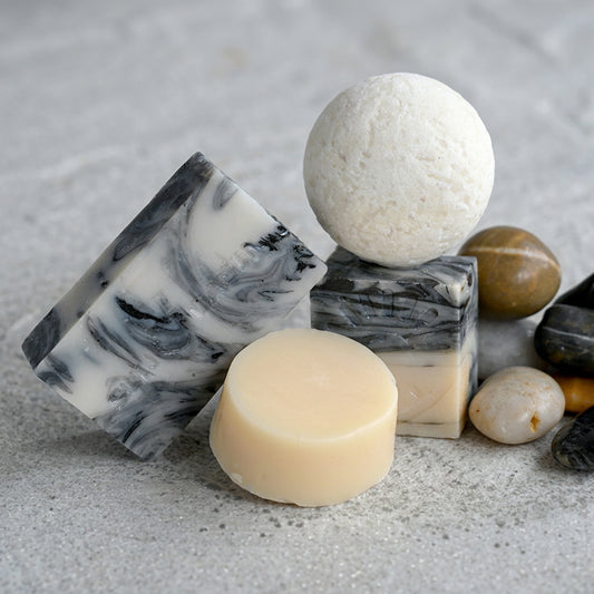 Four handmade bath products on a gray stone surface: a black-and-white marbled swirl soap bar, a matching marbled cube soap with an embossed logo, a round white bath bomb resting on top of the cube, and a small round cream-colored soap or lotion bar, surrounded by natural gray and tan stones.