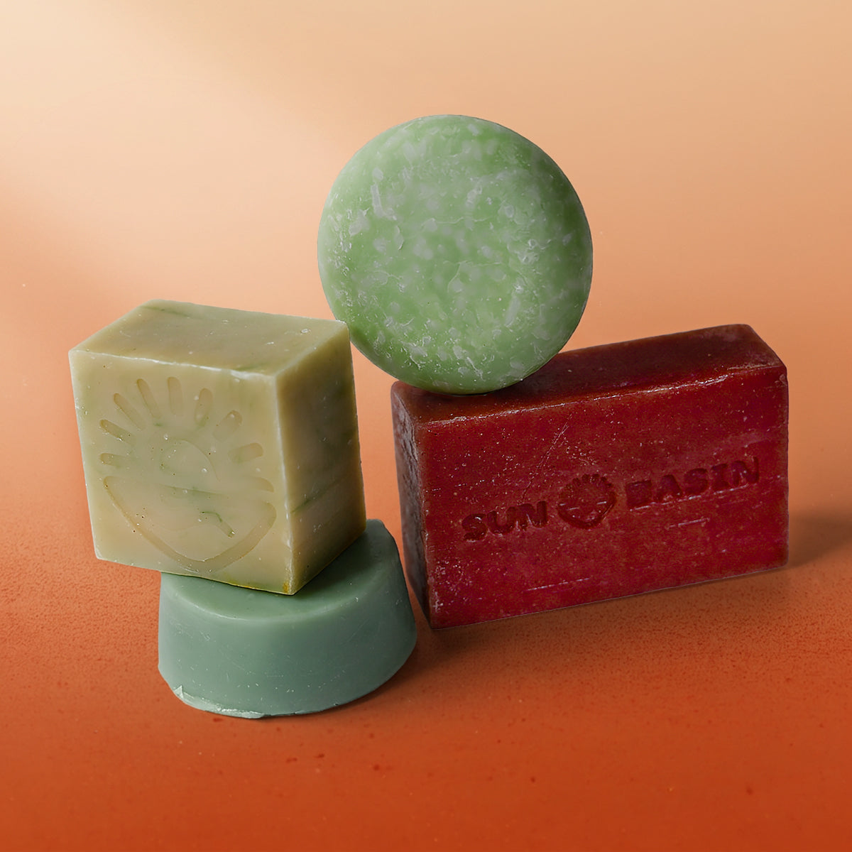 Four handmade soap bars on a warm orange background: a sage-green cube soap with an embossed sun and handprint design, a round green speckled soap, a smaller round teal soap, and a rectangular deep red bar stamped with a "Sun Basin" logo.