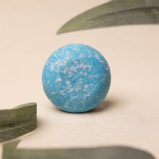 A round, textured light blue shampoo bar resting on a neutral beige surface with soft-focus green leaves in the background.
