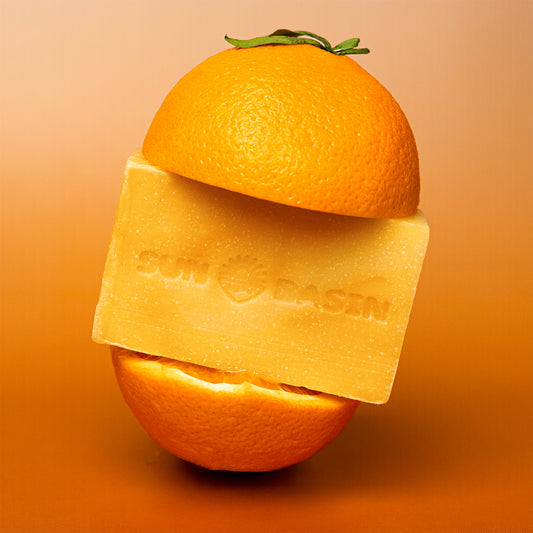 A rectangular bar of Sun Basin soap in a warm golden-orange color, positioned in front of a whole orange with its top half resting behind it. The soap bar is embossed with the brand name 'SUN BASIN' and a sun logo.