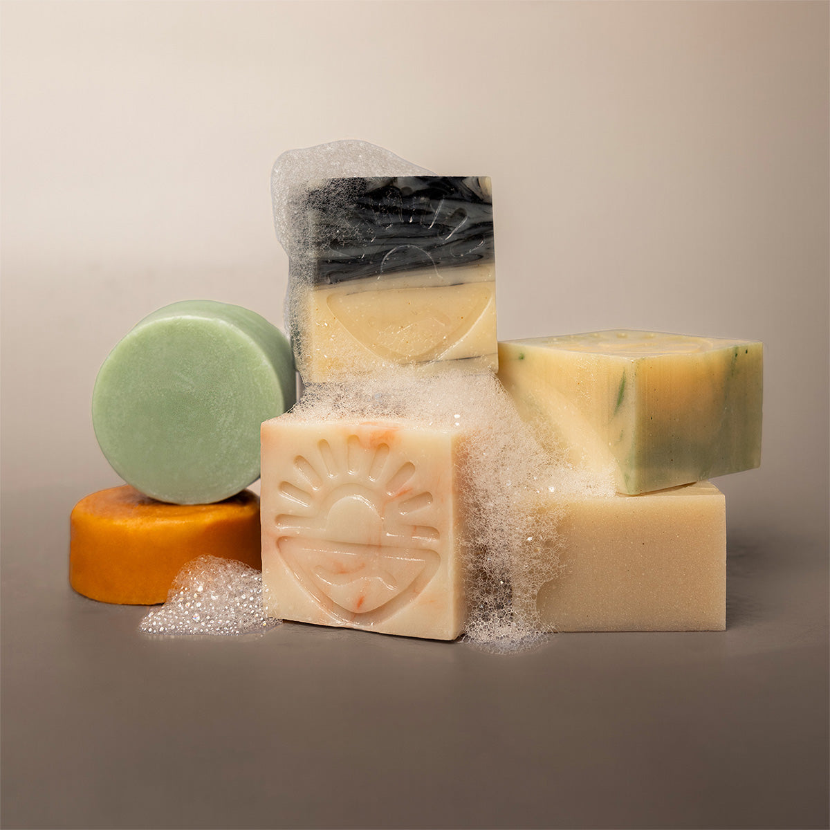 A group of artisanal handmade soaps in various shapes and colors, including a central cream soap with an embossed sun design, a green round soap, a marbled green and yellow soap block, and a black and yellow marbled soap wrapped in clear plastic, arranged on a neutral gray background with decorative soap bubbles.