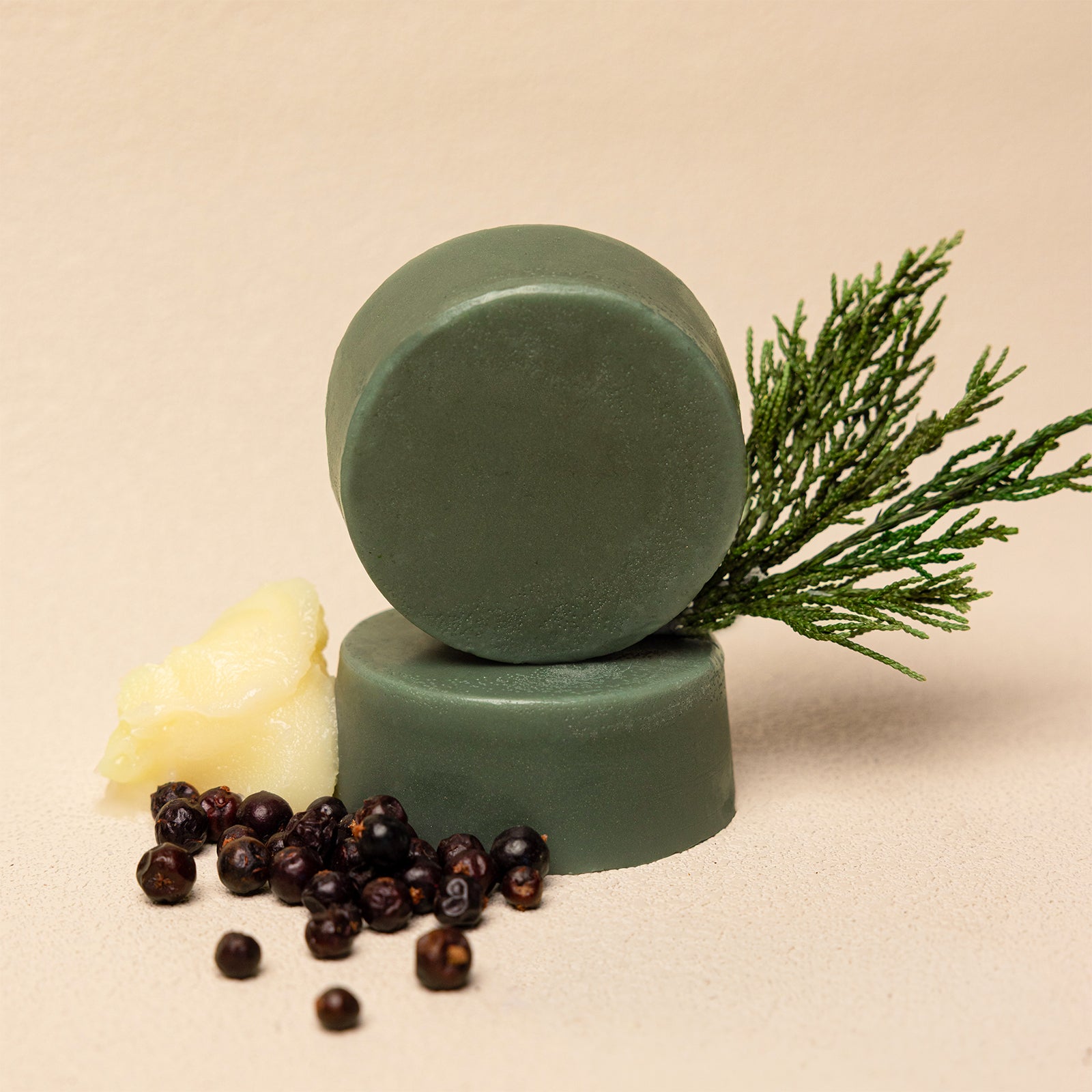 Two round green conditioner bars stacked on a beige background, styled with a piece of butter, dark whole spices, and a green pine branch.