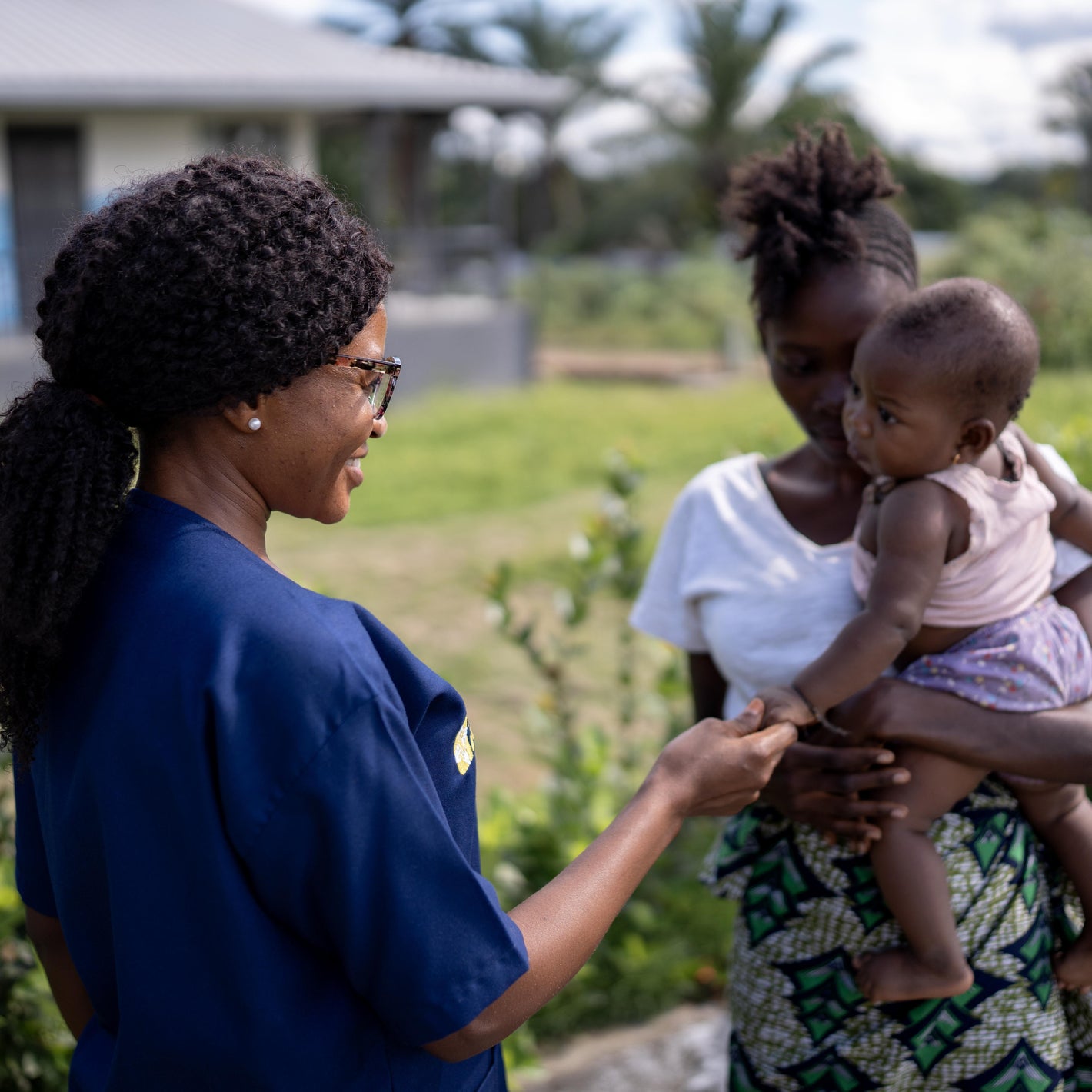 Woman in blue scrubs interacting with another woman holding a child outside of MCOE. Image provided by Partners In Health