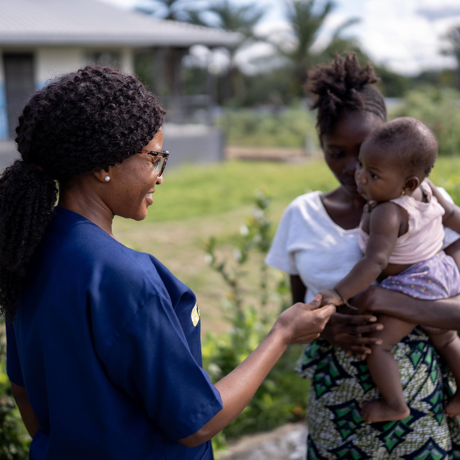 Woman in blue scrubs interacting with another woman holding a child outside of MCOE. Image provided by Partners In Health